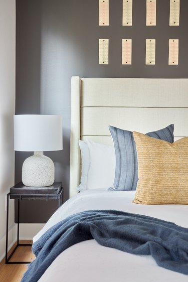Modern bedroom with off white headboard, white lamp, nightstand, blue blanket, pillows.